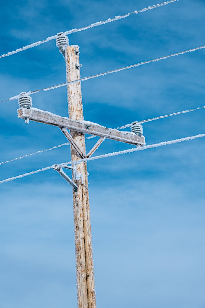 Wooden electric pole with snow-covered power lines against a blue sky.
